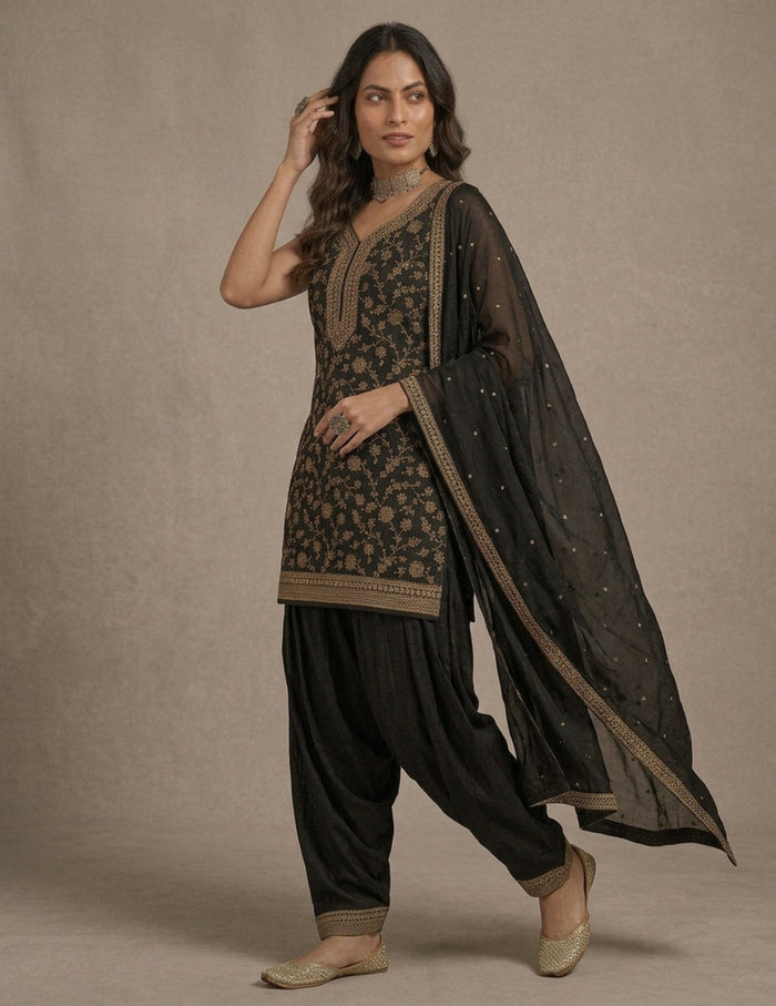 A woman modeling a black salwar kameez suit with intricate gold floral embroidery, black patola pants, and a sheer black dupatta with gold borders and small gold embellishments, set against a neutral beige studio backdrop.