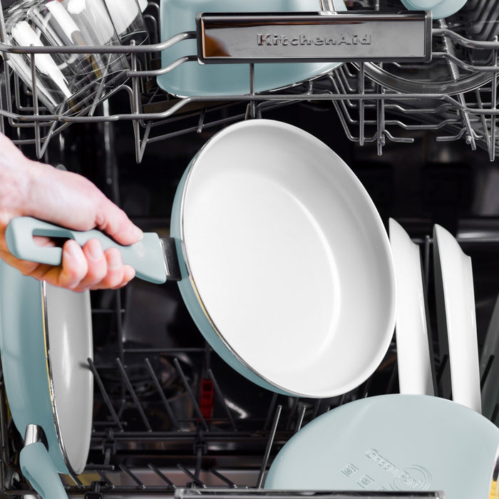 Blue ceramic nonstick frypan with spatula being loaded into a dishwasher alongside other similar cookware.