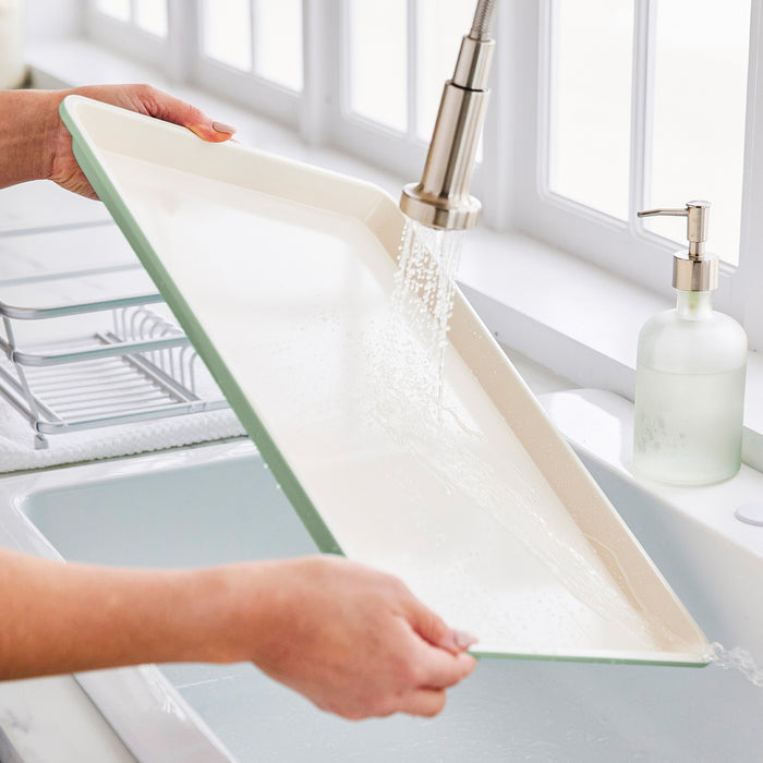 Sage green ceramic nonstick cookie sheet being rinsed under running water in a kitchen sink.