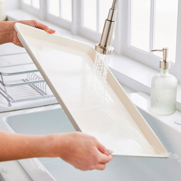 Person rinsing a GreenLife ceramic nonstick cookie sheet in a sunlit kitchen sink.