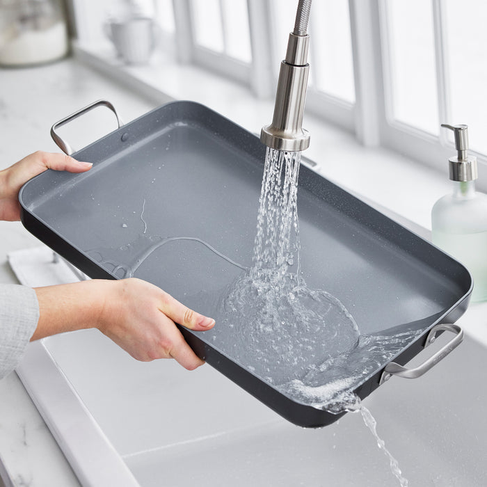 Washing a ceramic nonstick double burner griddle under a kitchen faucet in a bright sink.