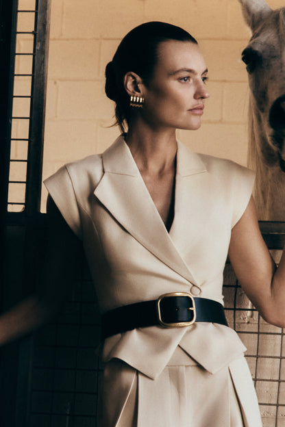 A model wearing a beige short-sleeve linen top with an asymmetric lapel neckline and a button closure. The top is paired with a mid-rise wide-leg linen pant.