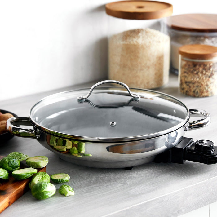 Electric skillet with a glass lid on a counter next to chopped Brussels sprouts and jars of grains.