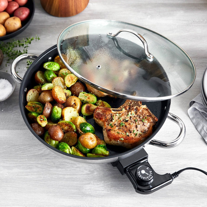 Electric skillet with glass lid cooking sautéed potatoes, Brussels sprouts, and a pork chop.