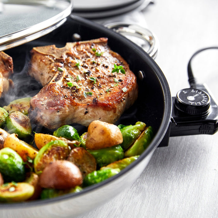 Electric skillet with glass lid cooking pork chops and mixed vegetables, topped with fresh herbs.