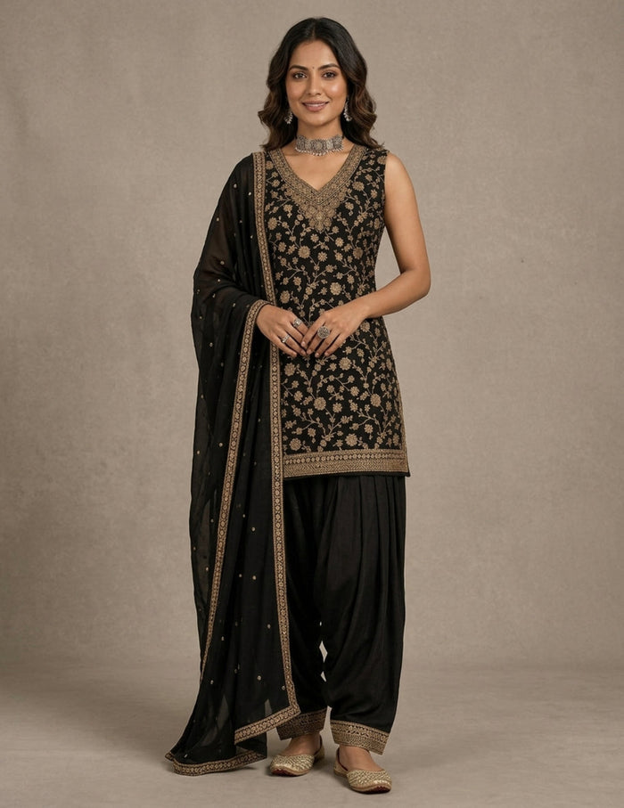 A woman models a traditional black Indian ethnic outfit with intricate gold embroidery, featuring a sleeveless embroidered kurta, black patola-style pants, and a matching black dupatta draped over her shoulder, set against a neutral beige studio background.