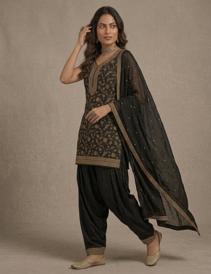 A woman modeling a black salwar kameez suit with intricate gold floral embroidery, black patola pants, and a sheer black dupatta with gold borders and small gold embellishments, set against a neutral beige studio backdrop.