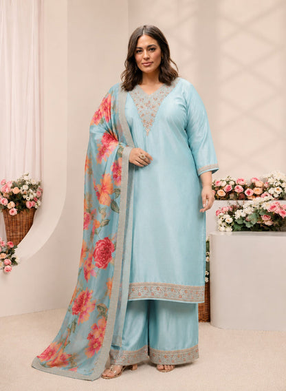 A woman modeling a light blue traditional Indian salwar kameez suit with a matching floral dupatta. The suit features intricate gold embroidery on the neckline, sleeve cuffs, and hem, and is styled with a sheer light blue dupatta adorned with large pink and red floral prints.