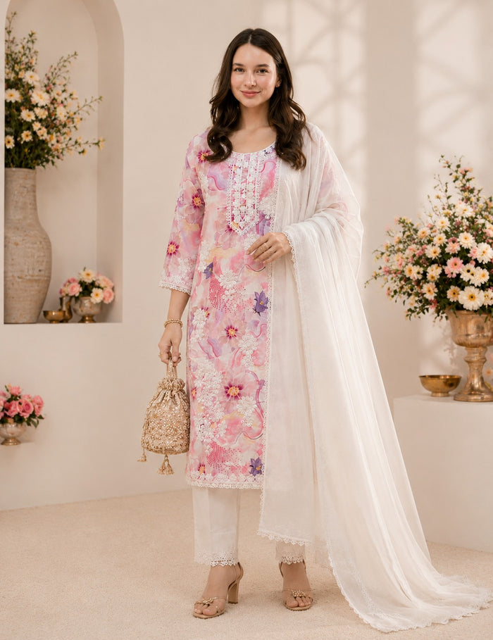 A woman models a three-piece traditional South Asian salwar kameez set. The set features a light pink embroidered kameez kurta with floral patterns, matching white trousers, and a sheer white dupatta.