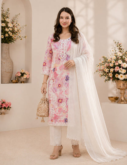 A woman models a three-piece traditional South Asian salwar kameez set. The set features a light pink embroidered kameez kurta with floral patterns, matching white trousers, and a sheer white dupatta.