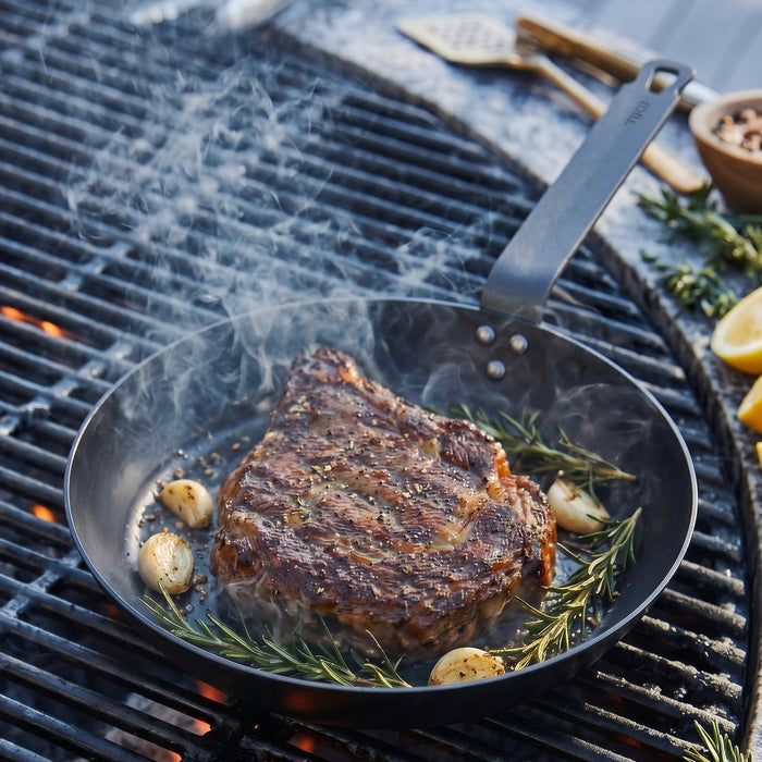 Steak with garlic and rosemary cooking in a carbon steel pan, smoke rising from the pan on a grill.