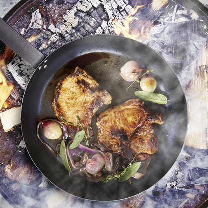 Pork chops and onions cooking in a black carbon steel pan from a 5-piece set.