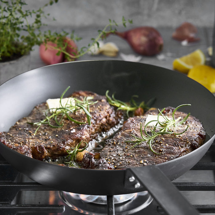 Two steaks with herbs and butter in a 12-inch carbon steel frypan, lemon and shallot beside the pan.