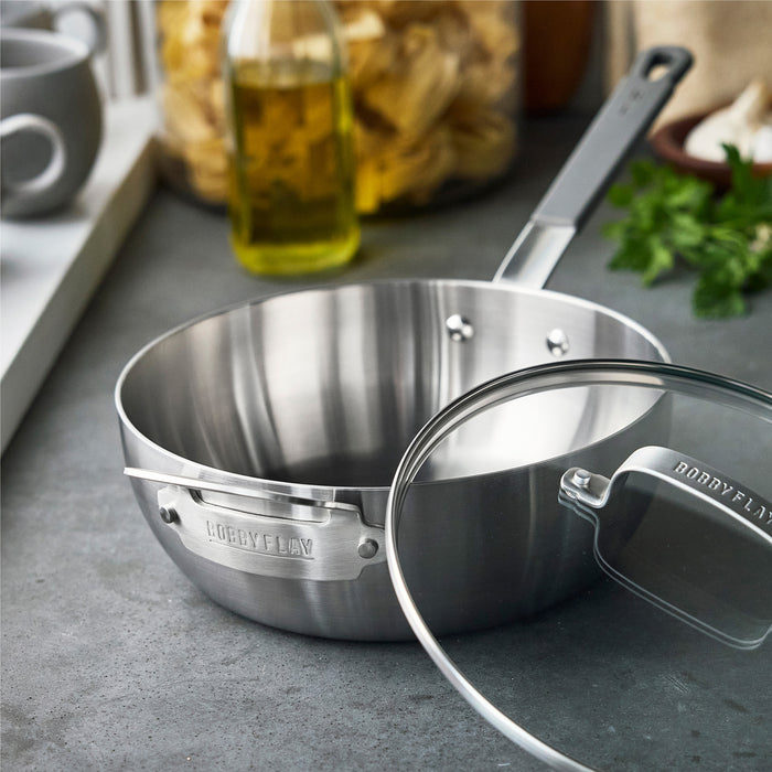 Stainless steel chefs pan with lid on a counter, olive oil and fresh herbs visible in the background.