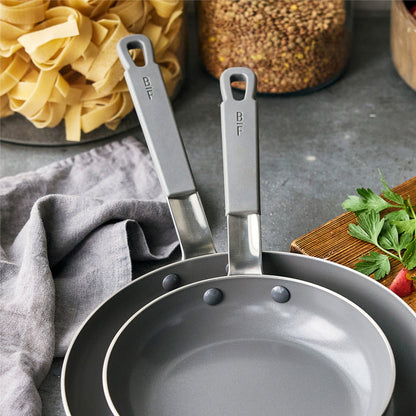 Set of two black nonstick pans with metal handles on a kitchen counter near herbs and jars.