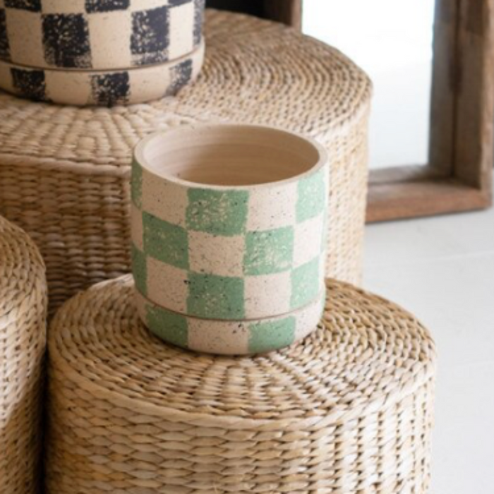 Three checkered ceramic plant pots in yellow, black-and-white, and mint green, displayed on a tiered stack of woven seagrass poufs with potted plants in a styled interior setting.