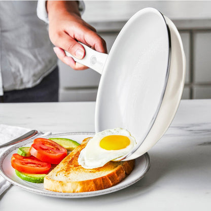 A hand uses a cream white ceramic nonstick frypan to slide a sunny-side-up egg onto avocado toast topped with tomato.