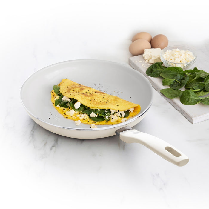 Cream white ceramic 10 frypan with a spinach and feta omelette, eggs, greens, and cheese on a wooden board.