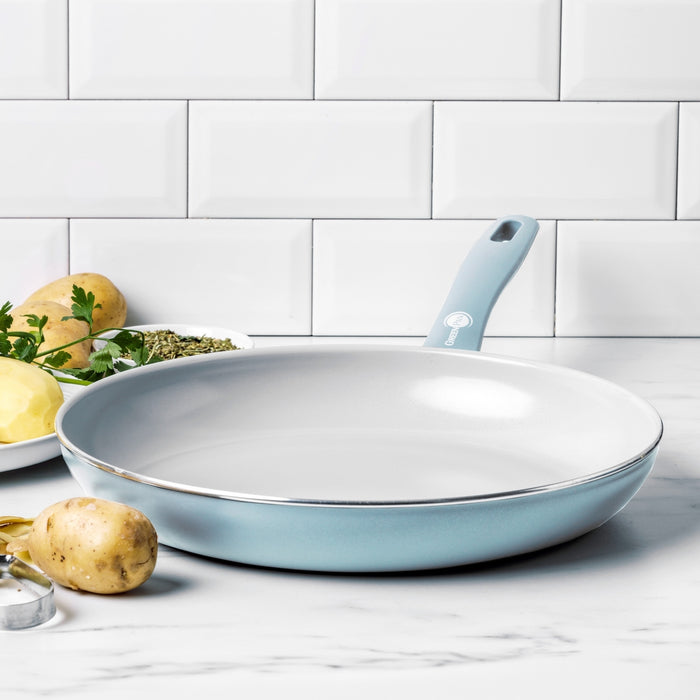 Blue haze ceramic nonstick 12-inch frypan on marble counter with potatoes, herbs, and white tiles in background.