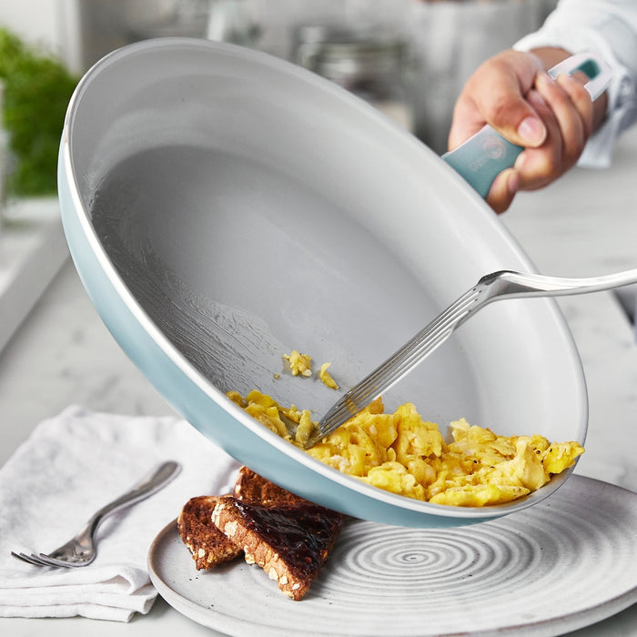 Scrambled eggs being served from a blue Dover ceramic nonstick frypan onto a plate with two granola bars.
