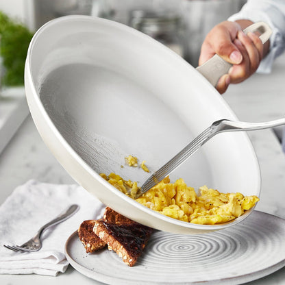Scrambled eggs being served from a cream white ceramic nonstick frypan onto a plate with toast.