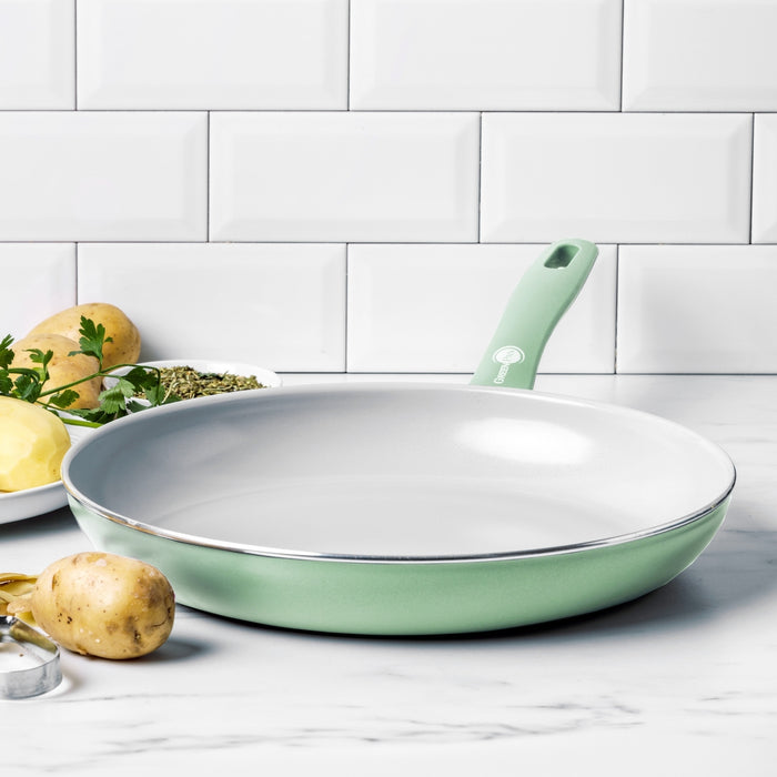 Pastel green ceramic nonstick frypan on marble counter with potatoes and herbs nearby.