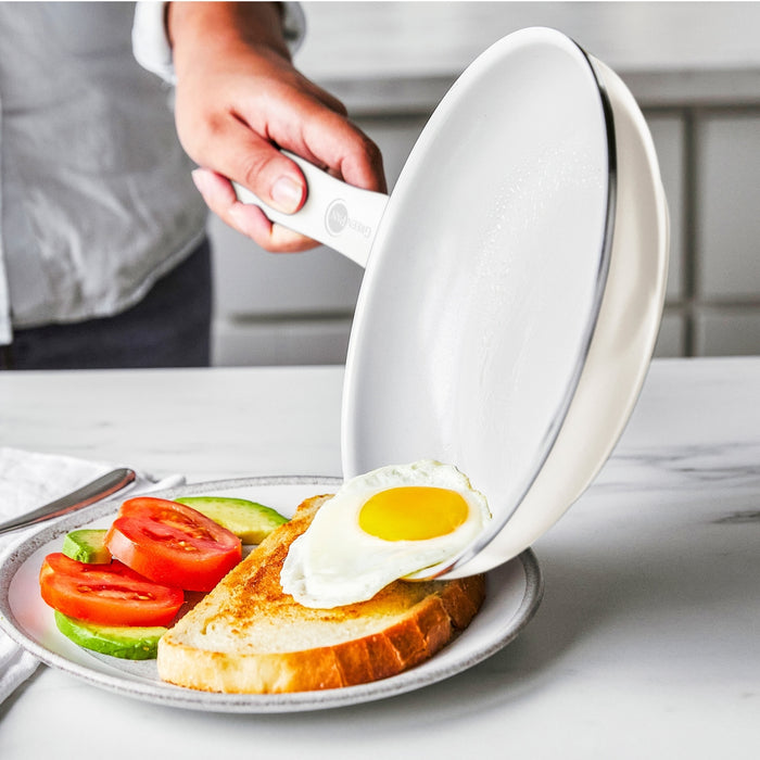 A hand slides a fried egg from a white ceramic nonstick pan onto toast with tomato and avocado slices.