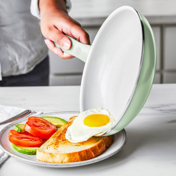 A hand slides a fried egg from a white ceramic nonstick pan onto toast with avocado and tomato on a plate.