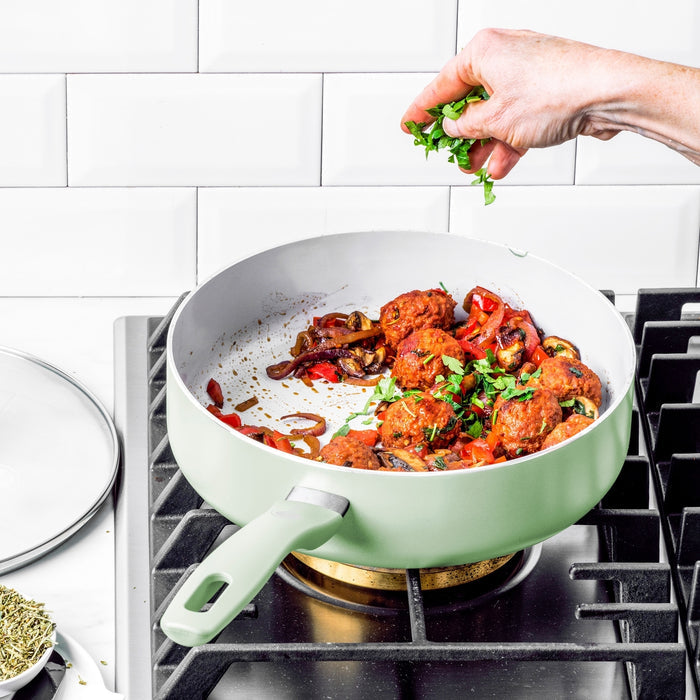 Hand sprinkles herbs over meatballs and vegetables in a pastel green ceramic cookware pan from a 12-piece set.