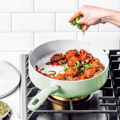 Hand sprinkles herbs over meatballs and vegetables in a pastel green ceramic cookware pan from a 12-piece set.