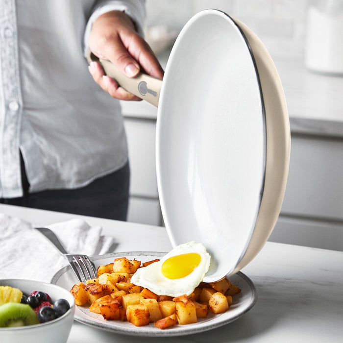 Hand sliding a fried egg from a white ceramic nonstick pan onto potatoes, with fruit on the kitchen counter.