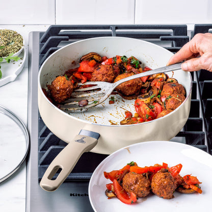 Hand serving meatballs with tomatoes and peppers from a black ceramic nonstick pan onto a white plate.