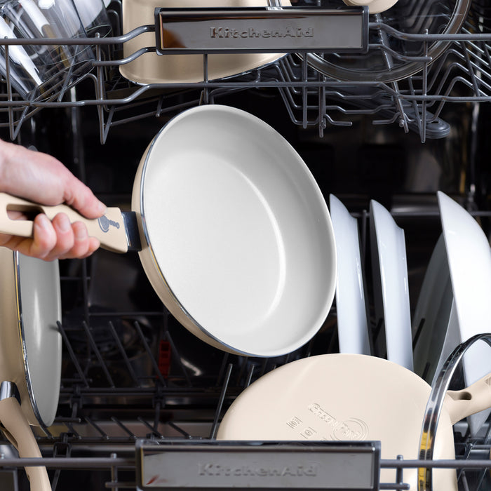 Hand placing taupe ceramic nonstick pan from 12-piece set into dishwasher with other dishes.
