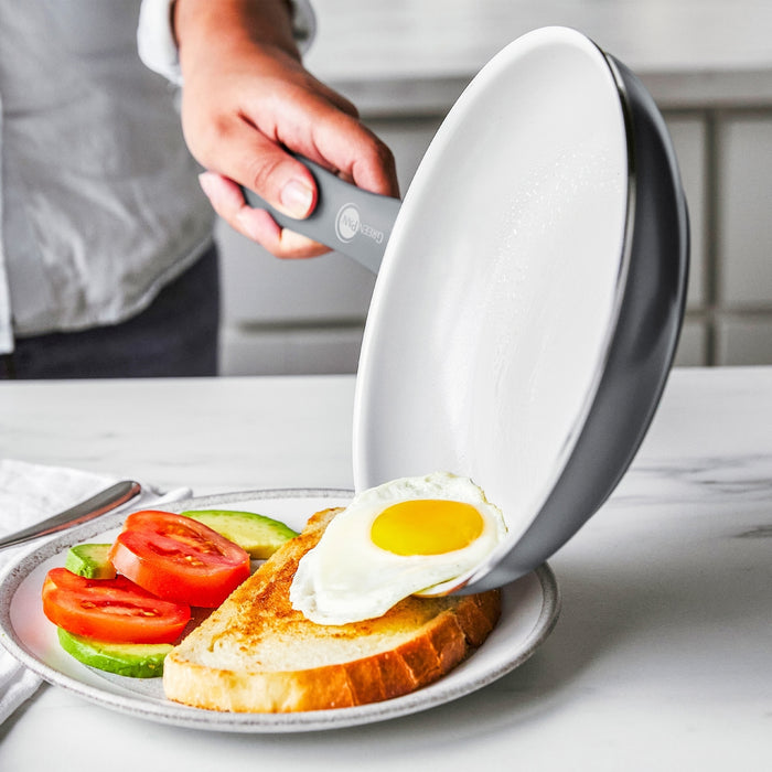 A fried egg slides from a white ceramic nonstick frypan onto toast, with tomato and avocado slices on the side.