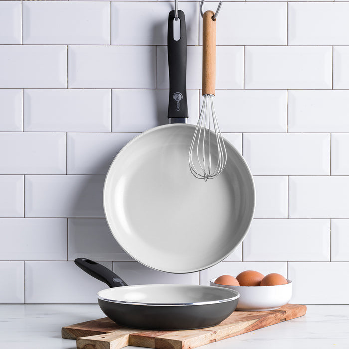 Black ceramic nonstick frypan set of two with a whisk and eggs on a wooden board in a tiled kitchen.