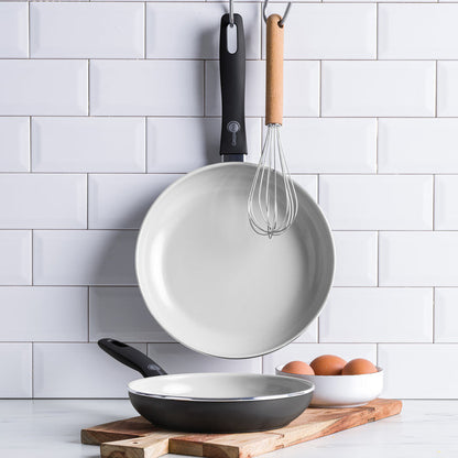 Black ceramic nonstick frypan set of two with a whisk and eggs on a wooden board in a tiled kitchen.