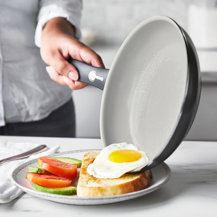 Fried egg being slid from a white ceramic nonstick frying pan onto toast with tomato and avocado.