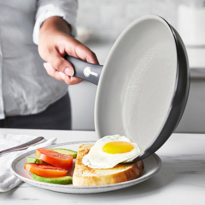Fried egg being slid from a white ceramic nonstick frying pan onto toast with tomato and avocado.