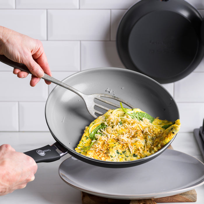A hand slides a cooked omelette from a gray ceramic nonstick frypan onto a white plate using a spatula.