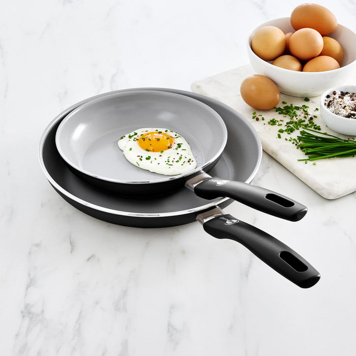 Black ceramic nonstick frypan set, 8 and 10, on marble countertop with eggs, chives, and spices nearby.