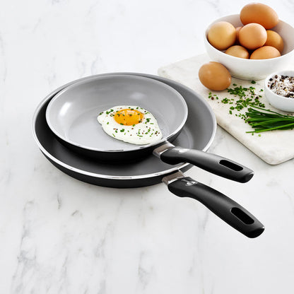 Black ceramic nonstick frypan set, 8 and 10, on marble countertop with eggs, chives, and spices nearby.
