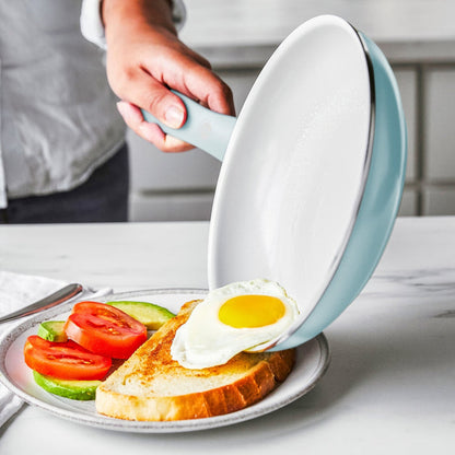 A hand slides a fried egg from a white ceramic nonstick frypan onto toast with tomato and avocado on a plate.