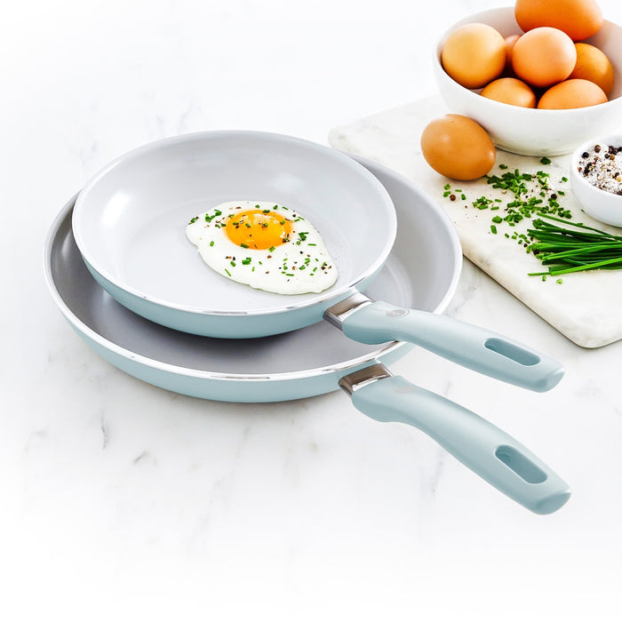 Blue ceramic nonstick frypan set, two pans with one holding a fried egg and fresh ingredients nearby.
