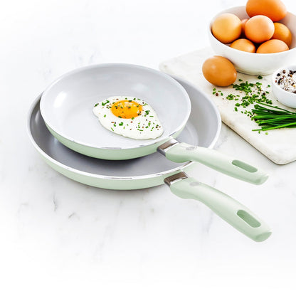 Two pastel green ceramic nonstick frypans with eggs, chopped chives, and salt on a white surface.