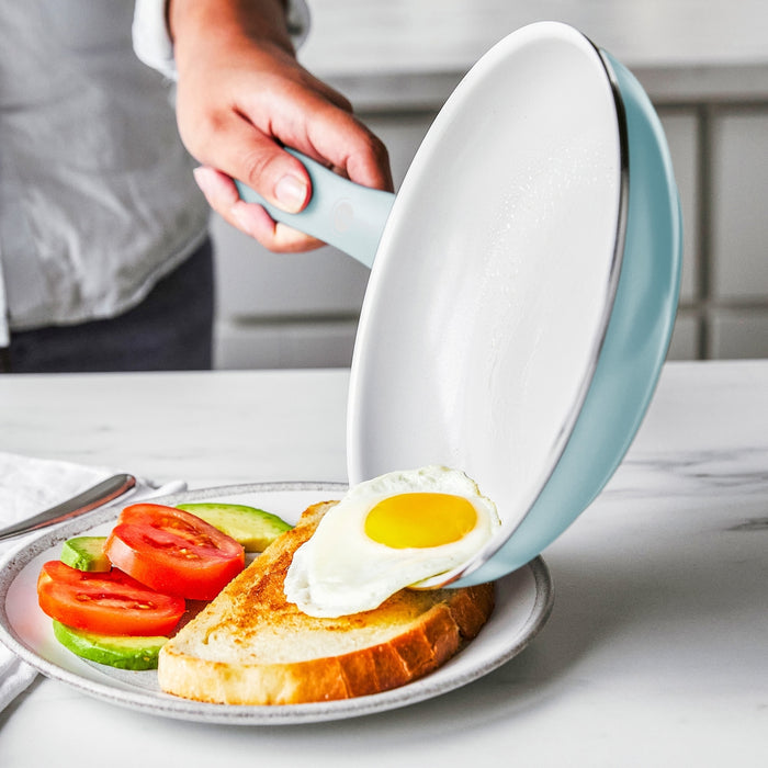 A hand slides a fried egg from a black ceramic nonstick frypan onto toast with tomato and avocado.