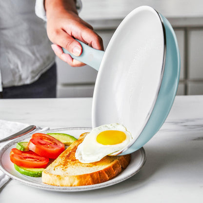 A hand slides a fried egg from a black ceramic nonstick frypan onto toast with tomato and avocado.