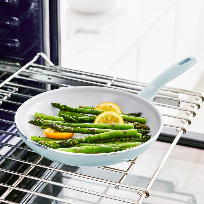 Blue ceramic nonstick frypan with spatula, holding asparagus and lemon, on a kitchen oven rack.