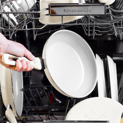 A cream white ceramic nonstick frypan with spatula being placed into a dishwasher next to white dishes.
