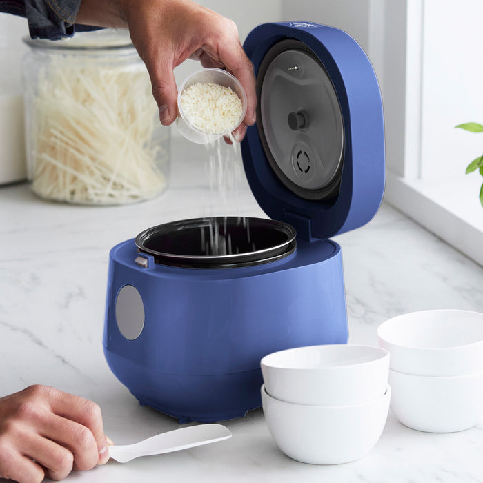 Rice being poured into a blue GreenLife 4-cup rice cooker on a kitchen counter with white bowls nearby.