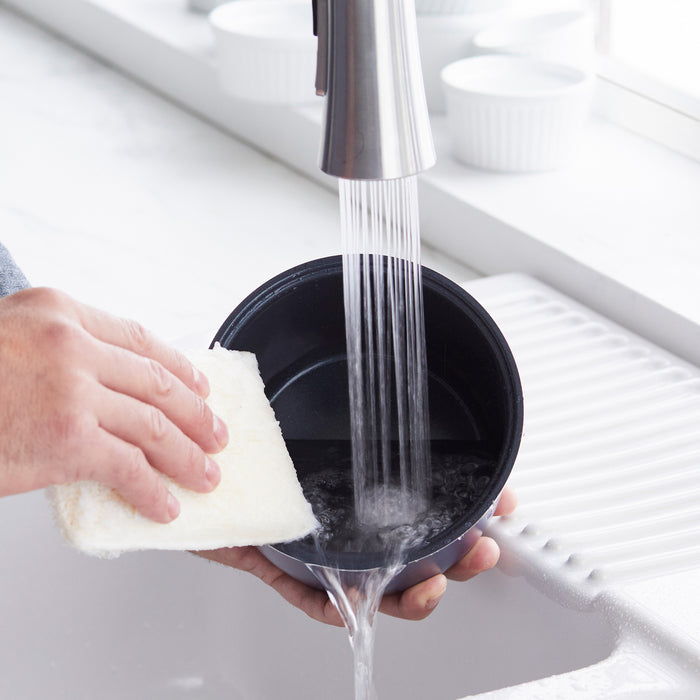 Hand washing a GreenLife rice cooker bowl under running water in a kitchen sink with a white sponge.